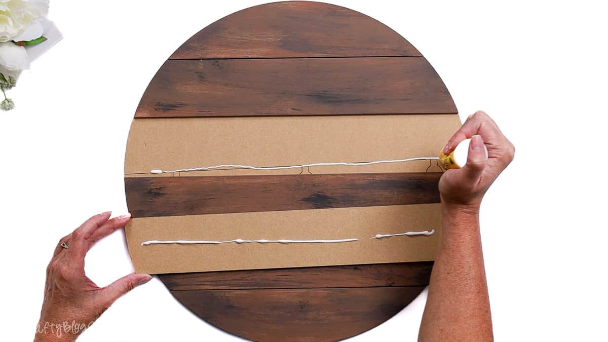 Hands applying wood glue to the back of a round shiplap wood door hanger, showing alternating painted and unpainted MDF slats.