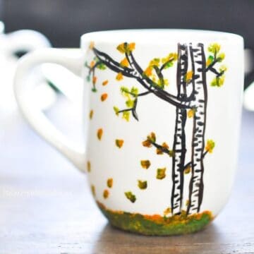 White ceramic mug hand-painted with birch trees, green and orange leaves, and a grassy base, sitting on a wooden table.