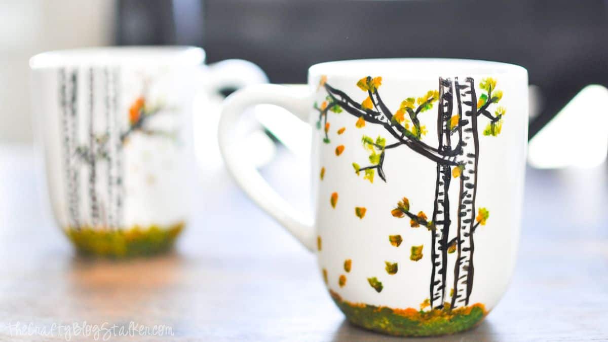 White ceramic mugs hand-painted with black birch trees and green and yellow leaves, sitting on a wooden table.