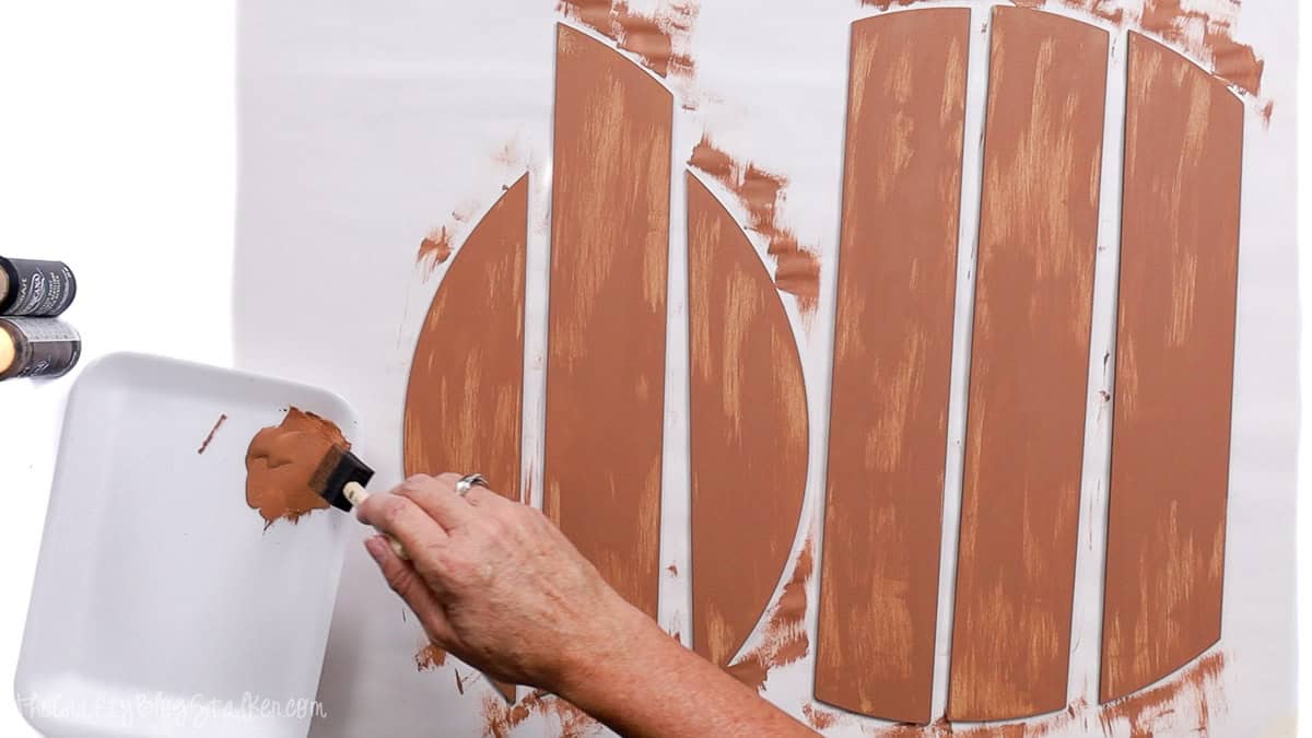 Hand painting MDF wood door hanger pieces with a foam brush and brown acrylic paint as the first base coat on a white work surface.