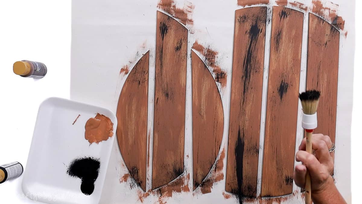 Hand dry brushing black paint over brown-painted MDF door hanger pieces to create a rustic wood grain effect on a white work surface.