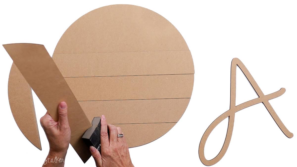 Hands sanding the edge of an MDF wood piece next to a round shiplap door hanger blank and a large unfinished wooden letter 