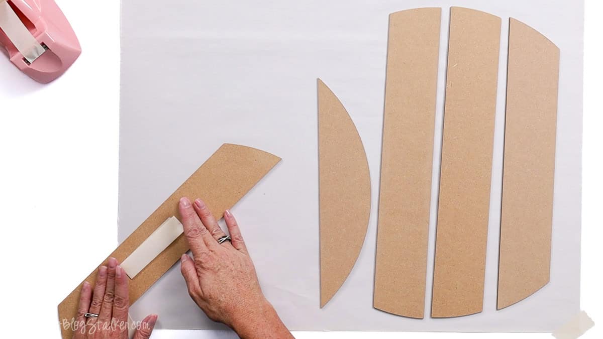 Hands applying tape to MDF wood pieces for assembling a round shiplap door hanger, with additional pieces laid out on a white surface.