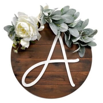 Finished round shiplap wood door hanger with a large white letter “A,” decorated with faux greenery and white flowers.