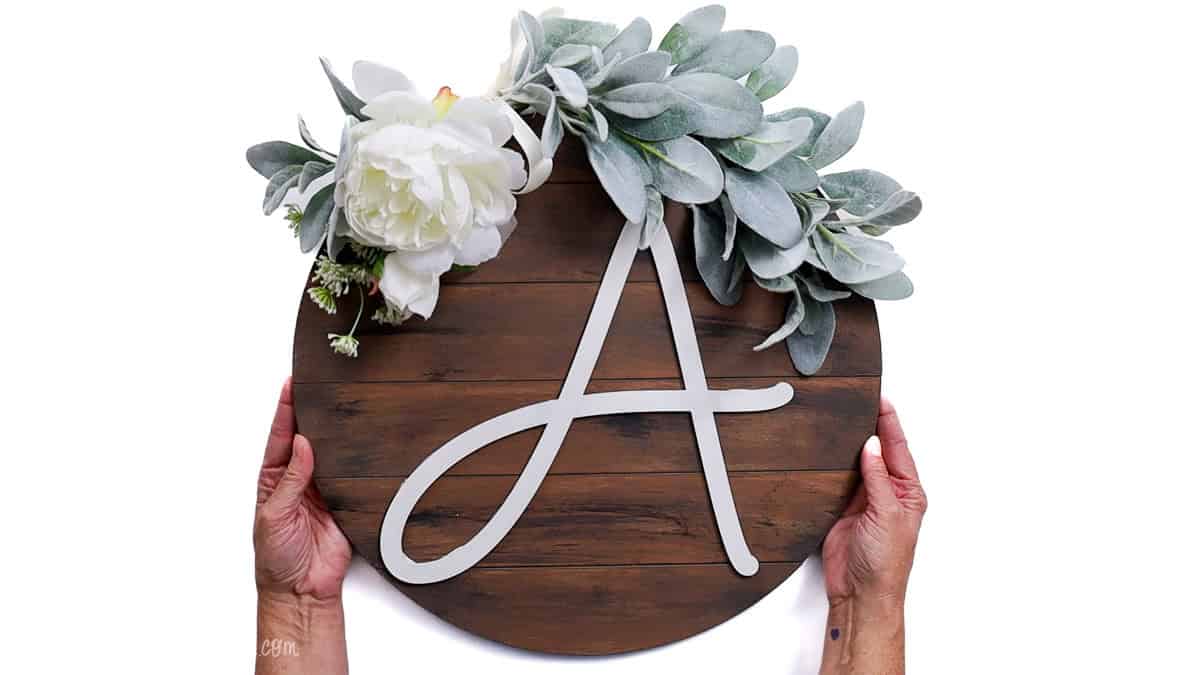 Hands holding a finished round shiplap wood door hanger decorated with faux greenery, white flowers, and a large white letter “A.”