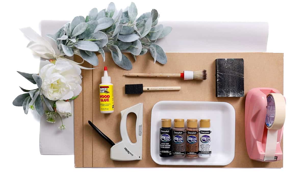 Flat lay of wood door hanger craft supplies including faux greenery, paint, brushes, wood glue, stapler, sandpaper, and masking tape.