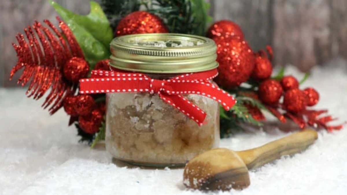 Jar of homemade cinnamon sugar body scrub tied with a red ribbon, surrounded by festive red ornaments and greenery on a snowy surface.