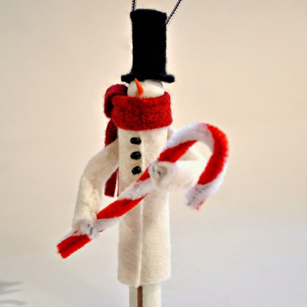 A handmade clothespin snowman ornament wearing a black top hat and red scarf, holding a red and white pipe cleaner candy cane.