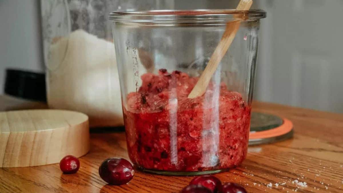 Glass jar filled with homemade cranberry sugar scrub and a wooden spoon, with fresh cranberries and sugar on a wooden table.