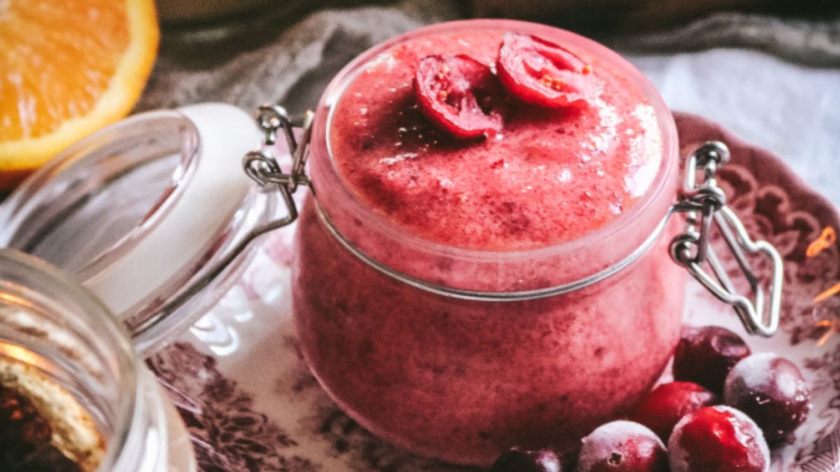 Jar of homemade cranberry sugar scrub topped with sliced cranberries, surrounded by fresh cranberries and an orange slice.
