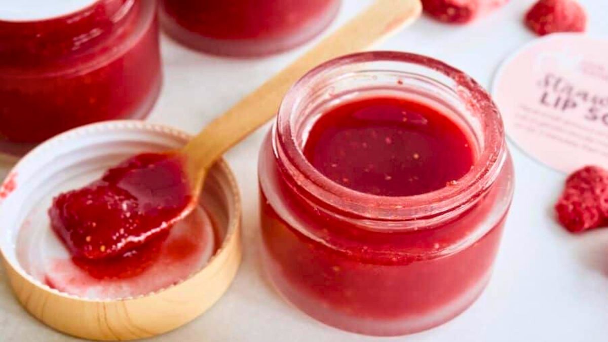 Jar of homemade strawberry lip scrub with a small wooden spoon resting on the lid, surrounded by pieces of dried strawberry.