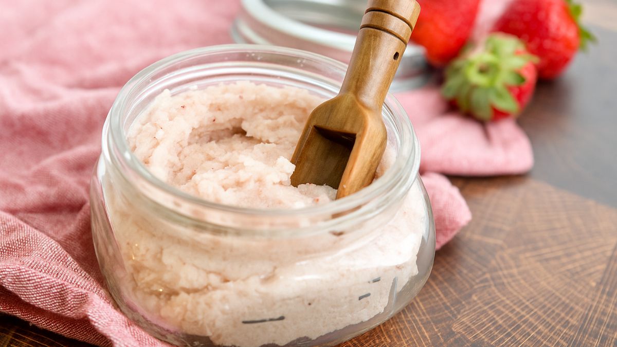 Jar of homemade strawberry sugar scrub with a small wooden scoop, placed on a pink cloth with fresh strawberries in the background.