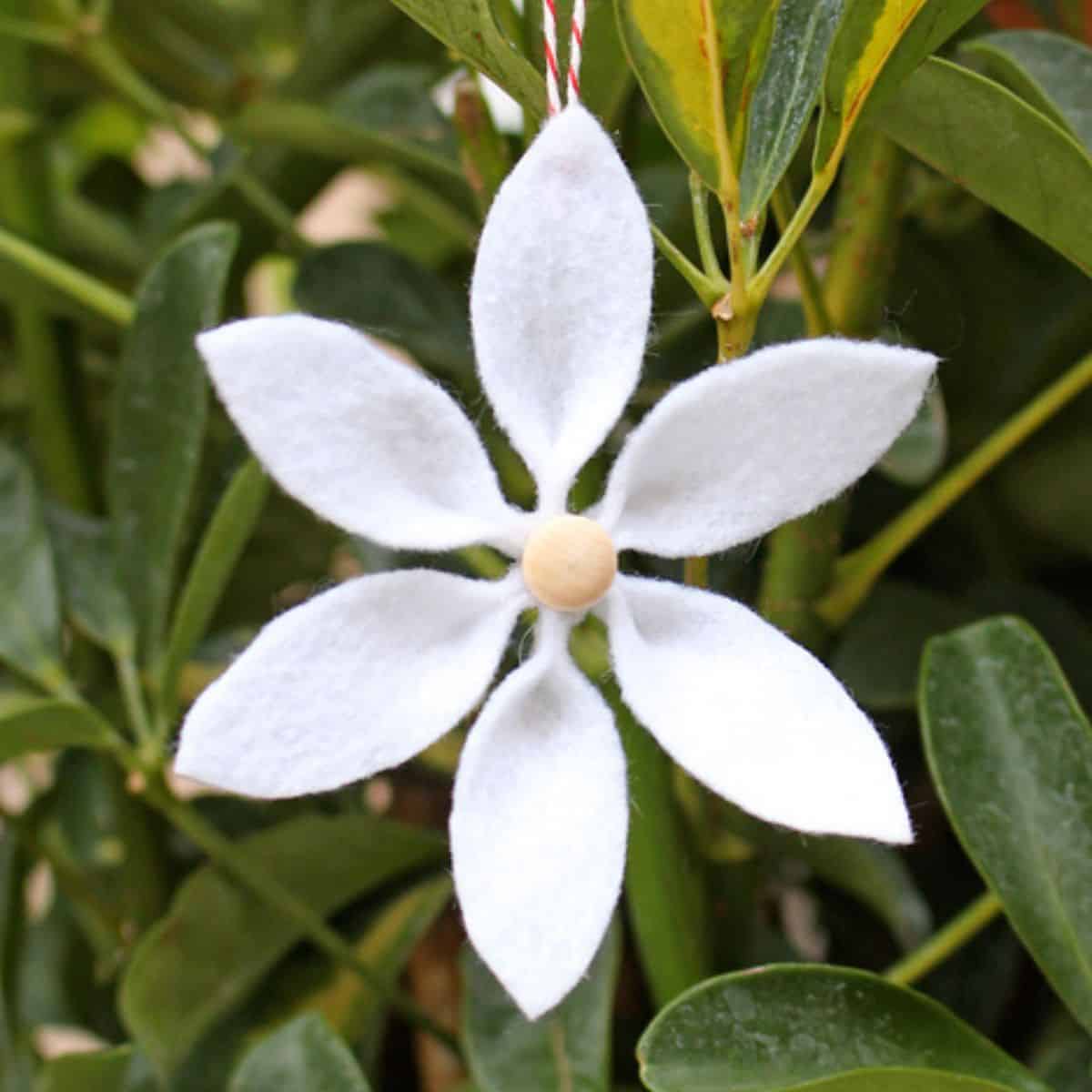 White felt flower ornament with six petals and a wooden bead center hanging in front of green leaves.