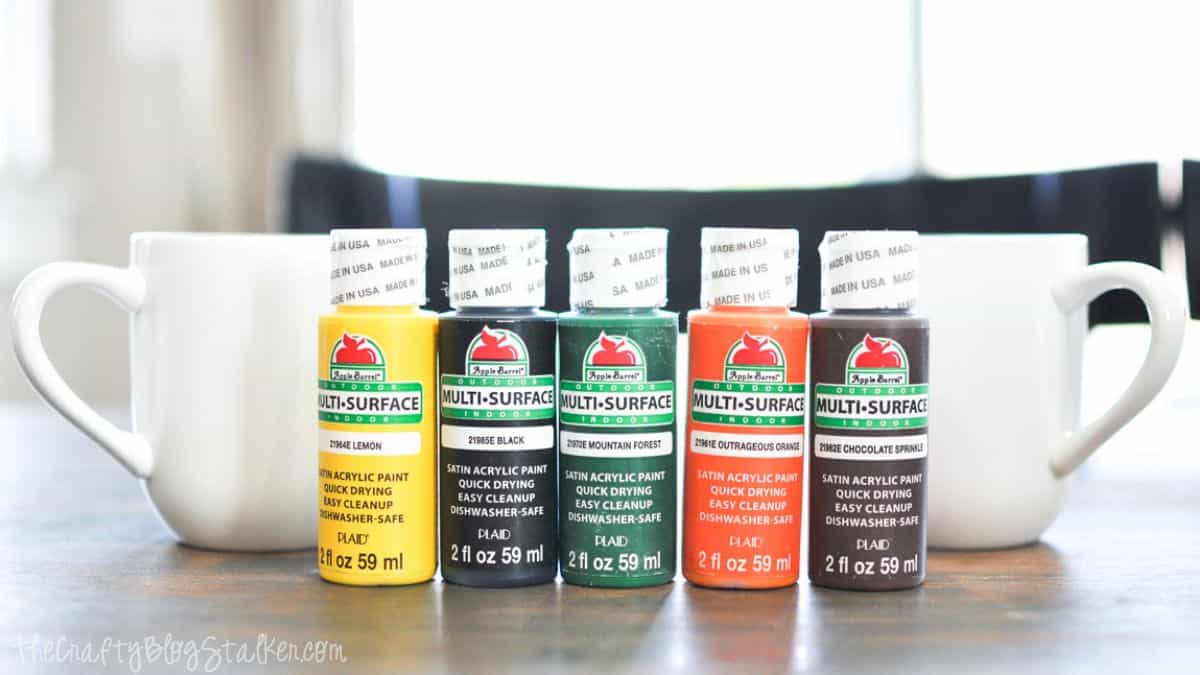 Five bottles of Apple Barrel multi-surface acrylic paint in assorted colors lined up between two plain white coffee mugs on a wooden table.