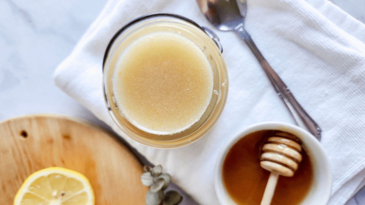 Jar of homemade honey lemon sugar scrub with a bowl of honey, spoon, and lemon slice on a white cloth background.