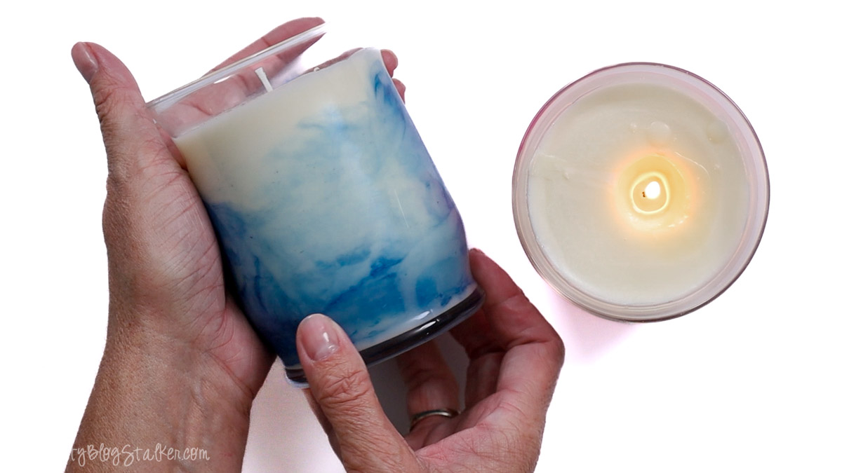 Hands holding a blue marbled candle in a glass jar next to a lit white candle, showing the finished look of homemade marbled soy candles.