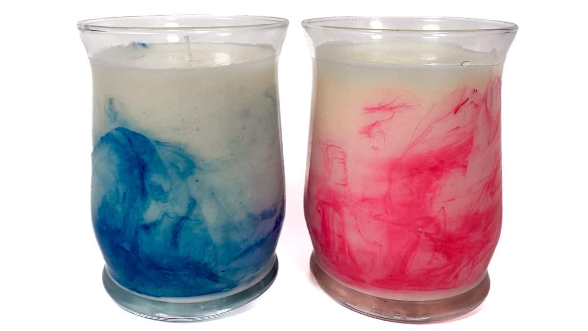 Two handmade candles in glass jars with colorful marbled designs, one in blue and one in pink, showing a DIY candle-making project.