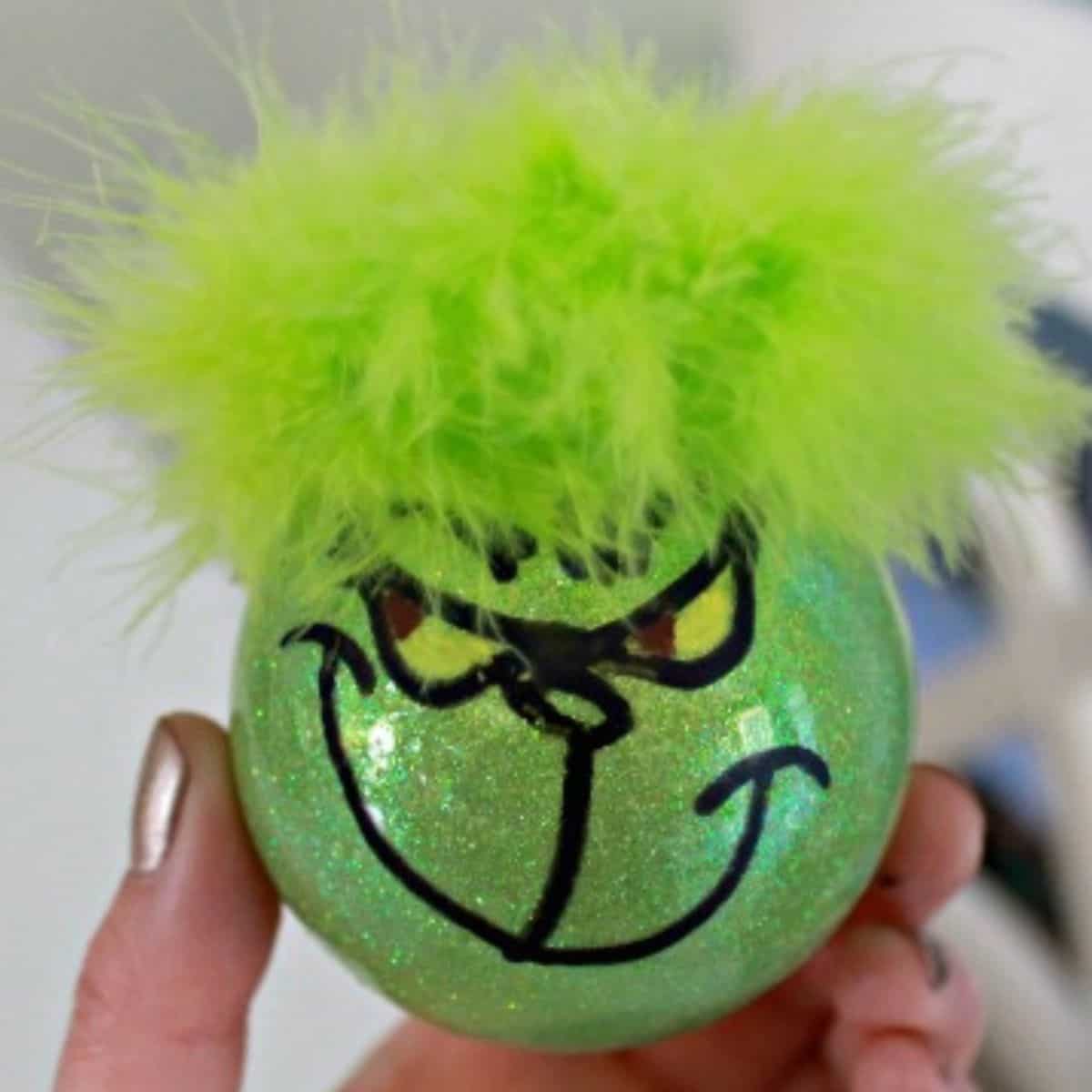 Hand holding a glittery green ornament painted with a Grinch face and topped with bright green fluffy feathers.