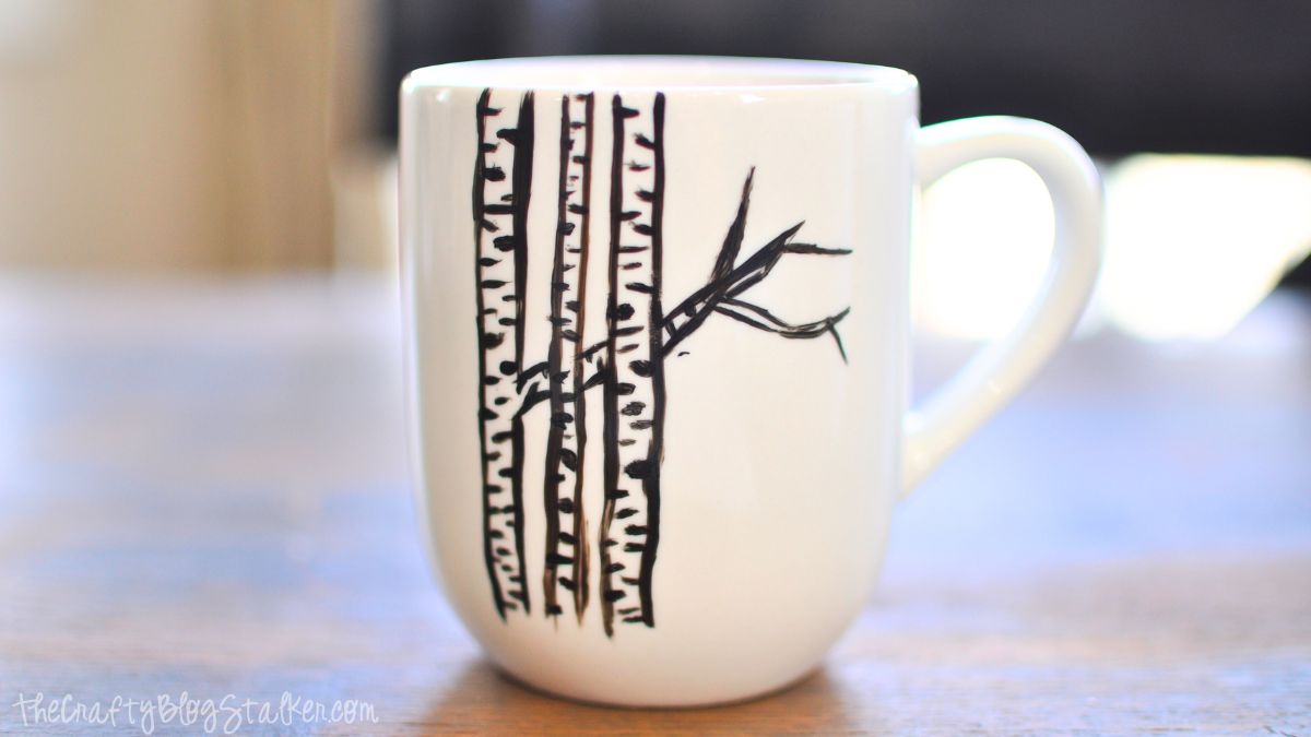 White coffee mug with a hand-painted black birch tree design featuring several trunks and branches on a wooden table.