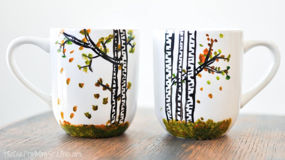 Two white coffee mugs hand-painted with black birch trees, green grass, and colorful autumn leaves on a wooden table.