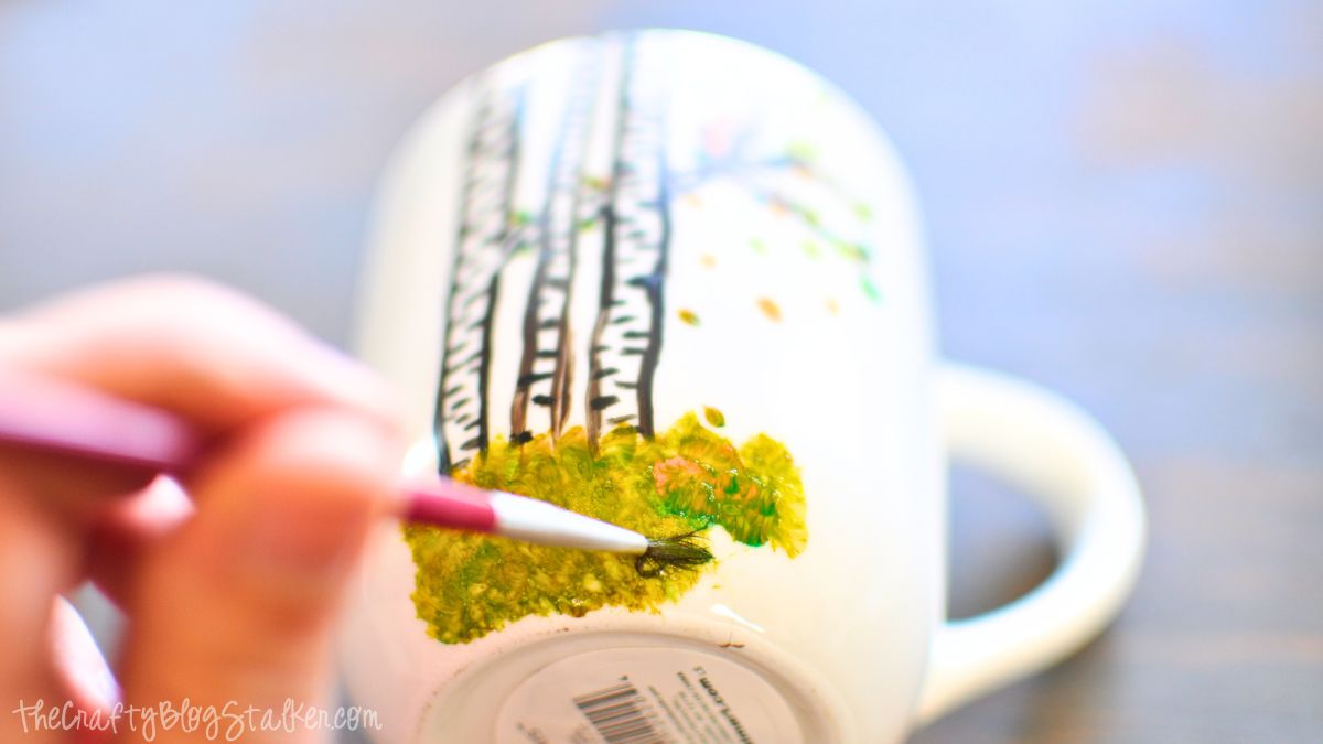A hand paints green leaves and grass details onto a white coffee mug featuring black birch tree designs.