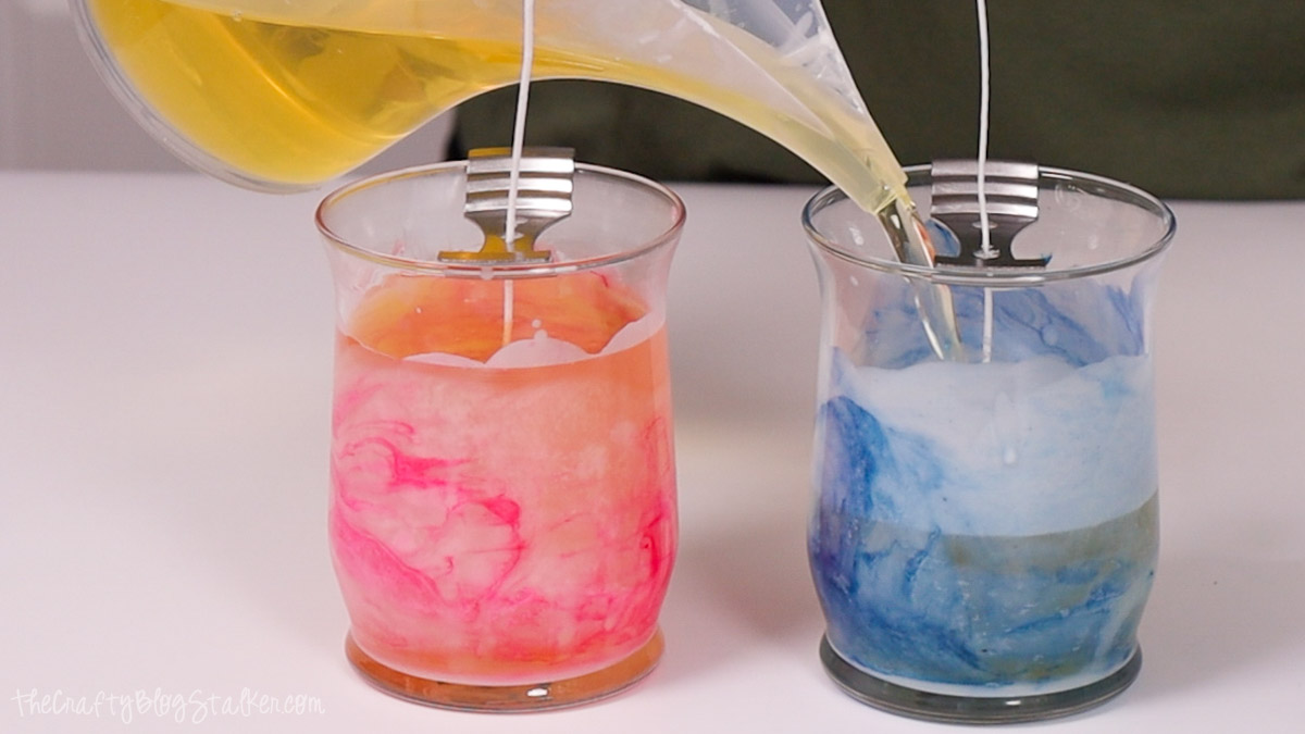 Pouring melted soy wax into glass jars with pink and blue marbled designs, each with a centered wick held by a metal wick holder for candle making.