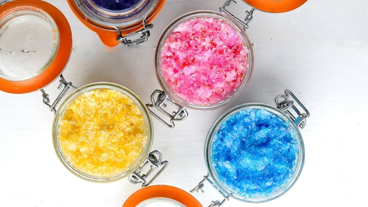 Colorful jars of homemade sugar scrubs in pink, blue, yellow, and purple with orange lids arranged on a white surface.