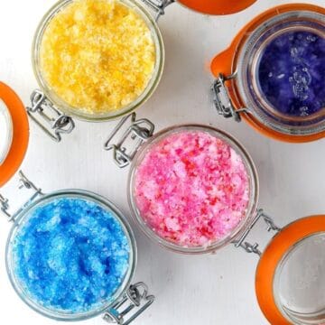 Colorful jars of homemade sugar scrubs in blue, pink, yellow, and purple arranged on a white surface with orange-lidded containers.