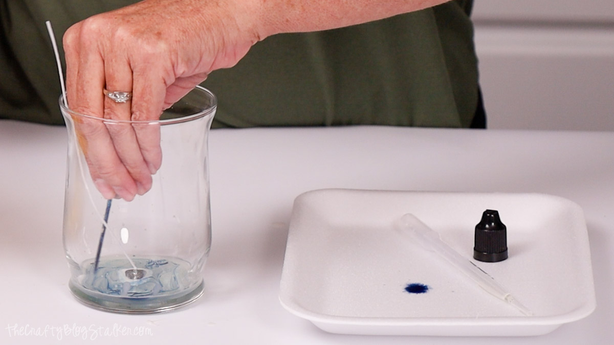 Person swirling blue candle dye inside a glass jar using a stick while preparing to make a handmade candle, with dye supplies on a tray nearby.