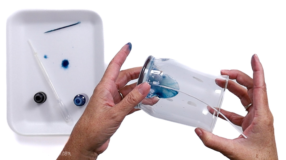 Person rotating a glass jar to spread blue candle dye inside evenly, creating a swirled marbled effect for a DIY candle project.