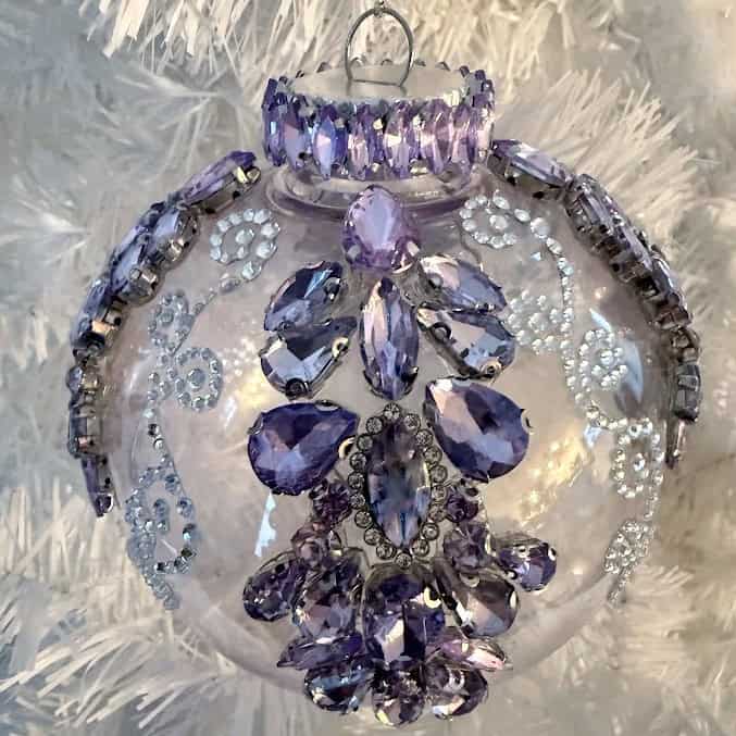 Clear glass Christmas ornament decorated with faceted rhinestones and crystal embellishments, hanging on a white artificial tree.