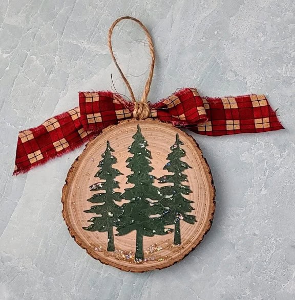 Round wood slice ornament with layered tree cutouts, twine hanger, and a plaid fabric bow, displayed on a textured surface.
