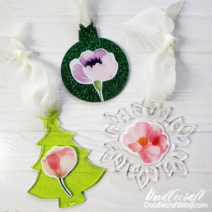 Three handmade floral ornaments with ribbon hangers, including a tree shape, a round ornament, and a snowflake, each decorated with a flower decal.