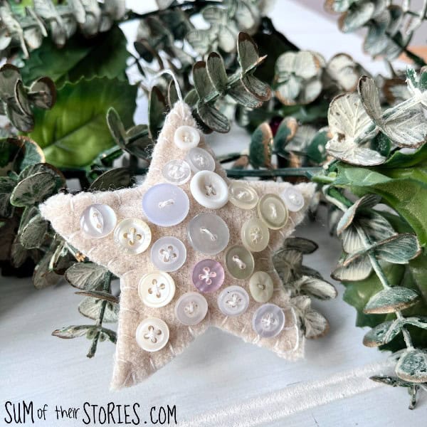 Handmade star-shaped ornament covered with assorted buttons, hanging among greenery on a white surface.