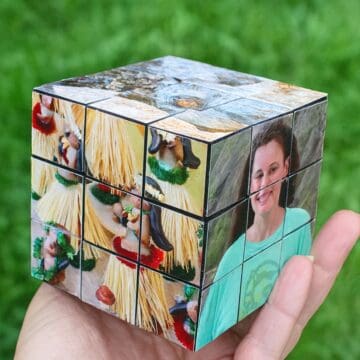 Hand holding a personalized photo cube with small square panels forming a Rubik’s cube style design, featuring family photos against a green grass background.