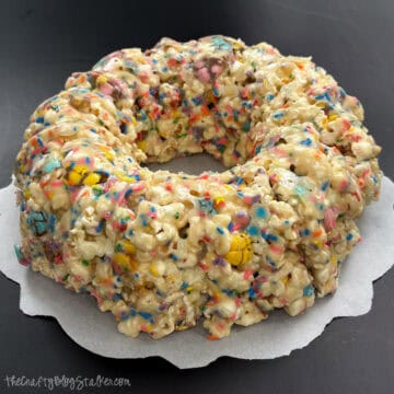 Ring-shaped popcorn cake made with marshmallow-coated popcorn and candy sprinkles, formed like a bundt cake and set on parchment paper.