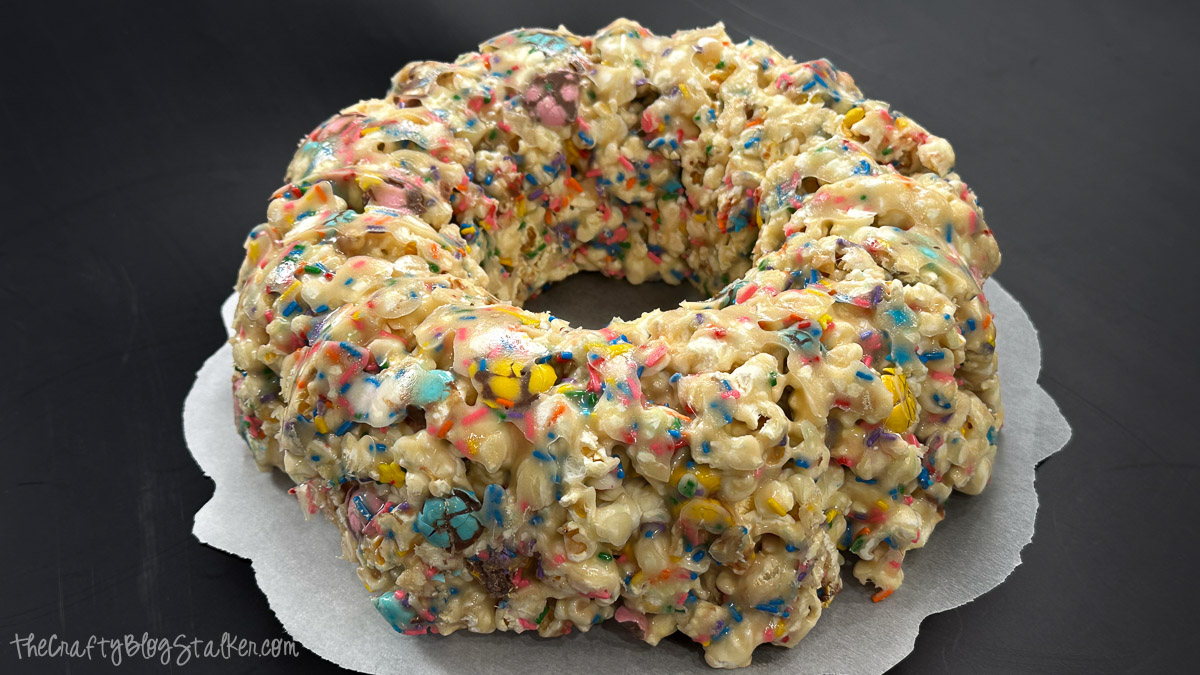 Ring-shaped popcorn cake made from marshmallow-coated popcorn with candy pieces and sprinkles, displayed on parchment paper against a dark background.