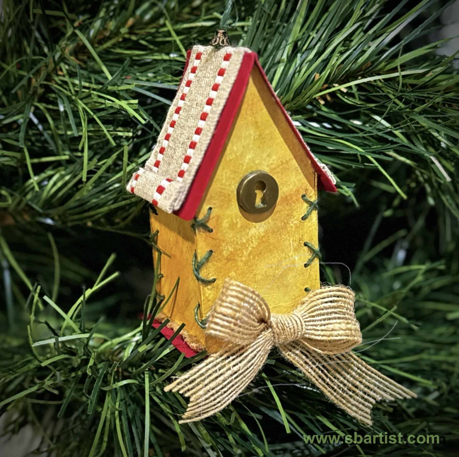 Handmade paper birdhouse ornament hanging on a Christmas tree, decorated with stitched details, a ribbon bow, and a button accent on the front.