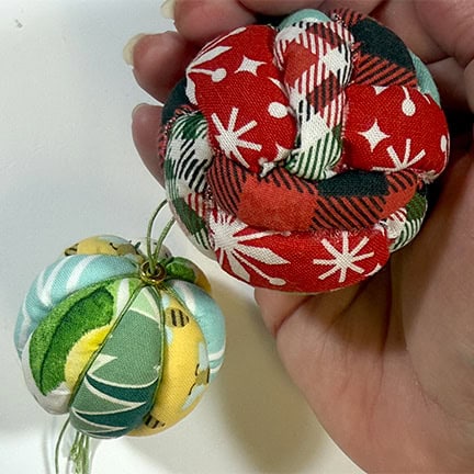 Two handmade fabric ornaments made from patterned fabric, one ball-shaped and one knot-style, held in a hand to show their size and texture.