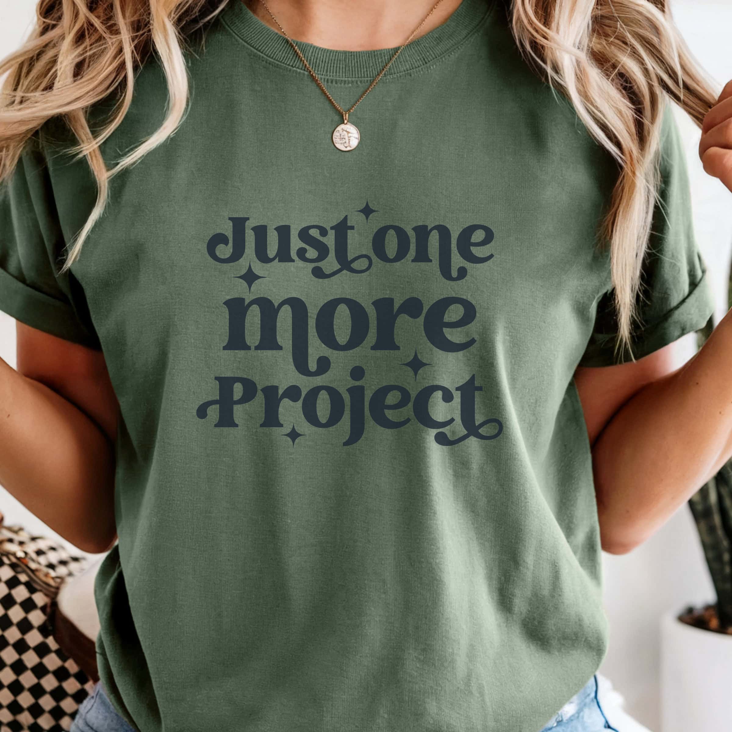 Person wearing a T-shirt with the phrase Just one more project printed on the front, styled with casual jeans and a necklace.