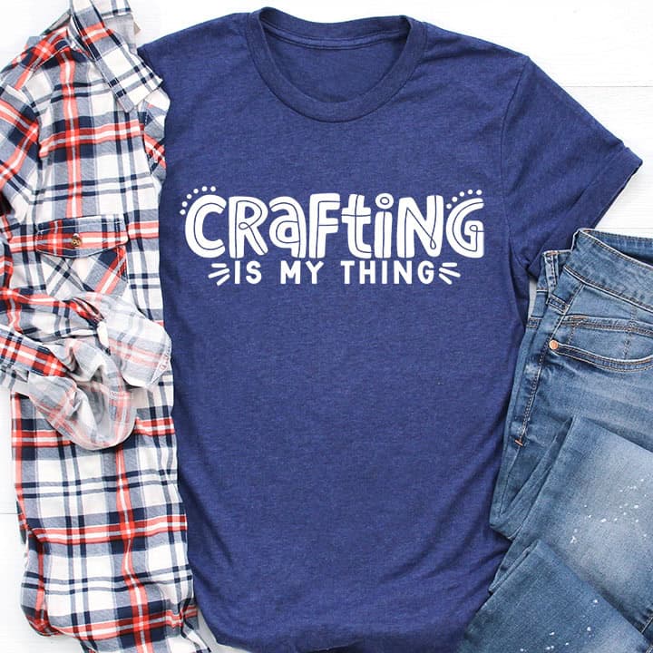 Flat lay of a T-shirt reading Crafting Is My Thing, styled with a plaid shirt and jeans on a light surface.
