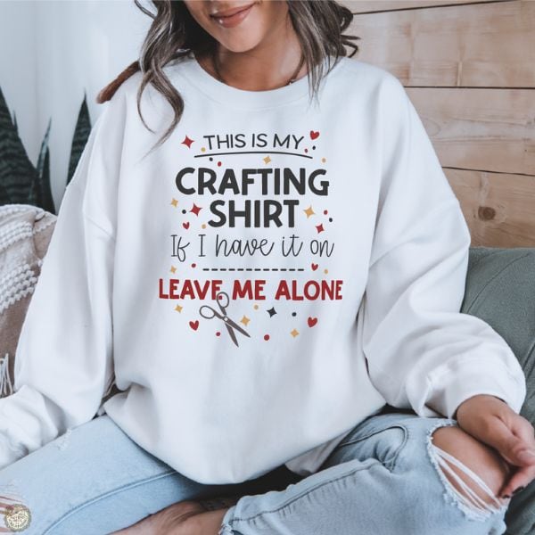 Person sitting indoors wearing a sweatshirt that reads This Is My Crafting Shirt If I Have It On Leave Me Alone, with decorative icons around the text.