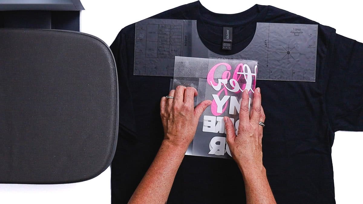 Hands positioning an iron-on vinyl design on a T-shirt using alignment guides, with a heat press platen nearby before pressing.