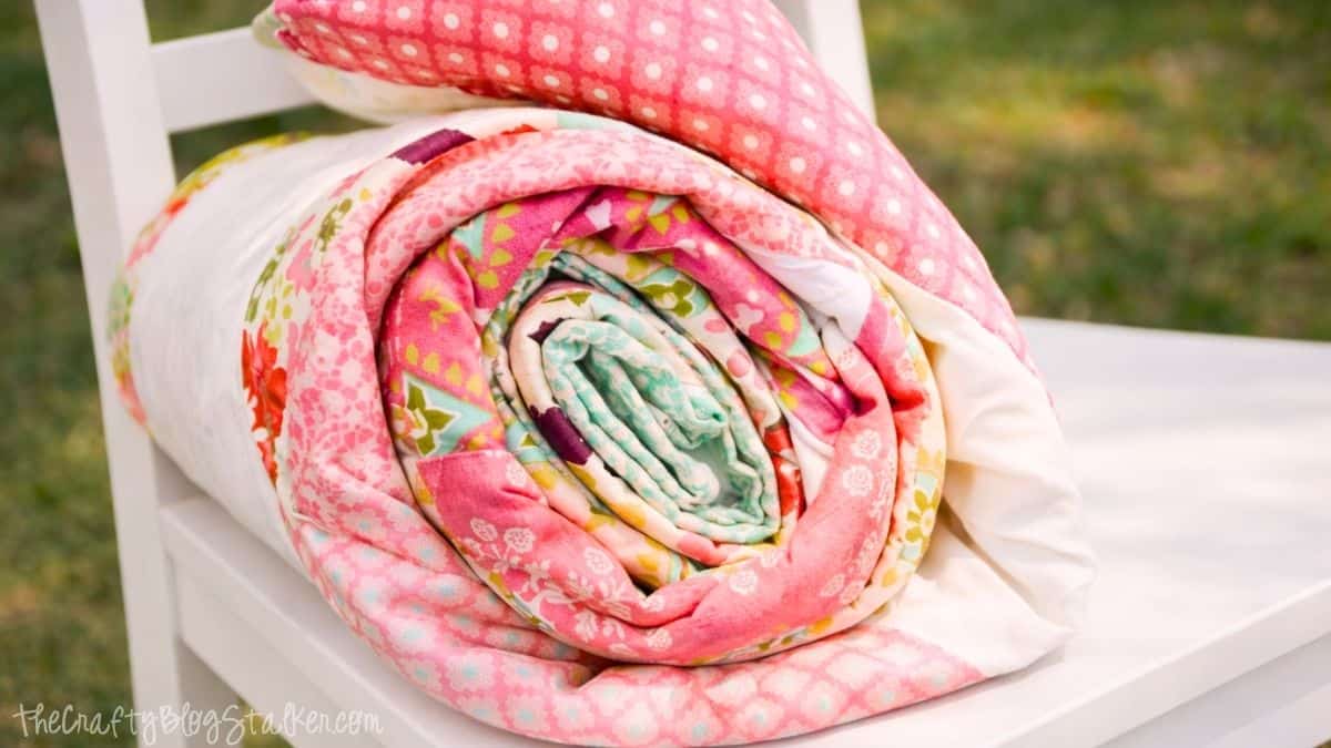 Rolled handmade quilt resting on a white chair outdoors, showing layered patchwork fabric with a zig zag design.
