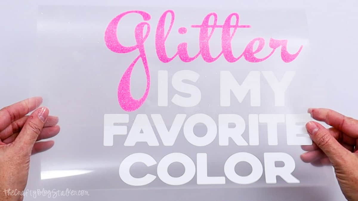 Hands holding a weeded iron-on vinyl design that reads Glitter Is My Favorite Color, with the cut lettering attached to a clear carrier sheet.