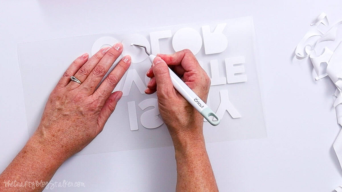 Hands using a weeding tool to remove excess iron-on vinyl from a cut text design laid on a flat work surface.