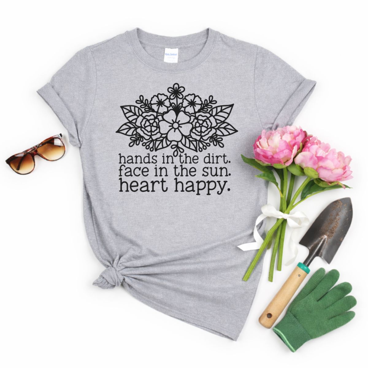 Gray T-shirt with floral graphic and the phrase hands in the dirt face in the sun heart happy, styled with sunglasses, pink flowers, gloves, and a garden trowel.