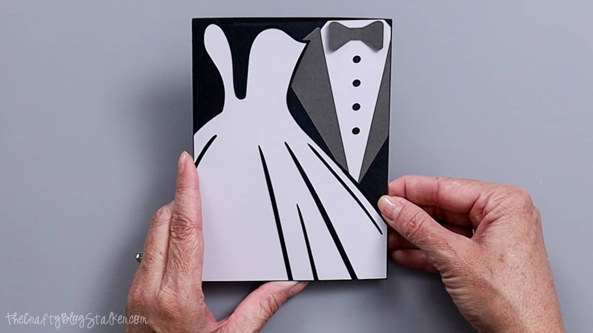 Hands holding a handmade wedding card featuring a white bride's dress and groom's tuxedo design cut from layered cardstock.