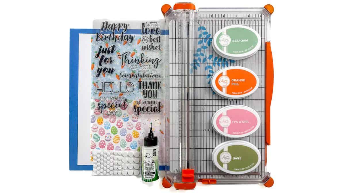 Cardmaking supplies including patterned paper, clear sentiment stamps, ink pads, paper trimmer, glue, and foam dots arranged on a white background.