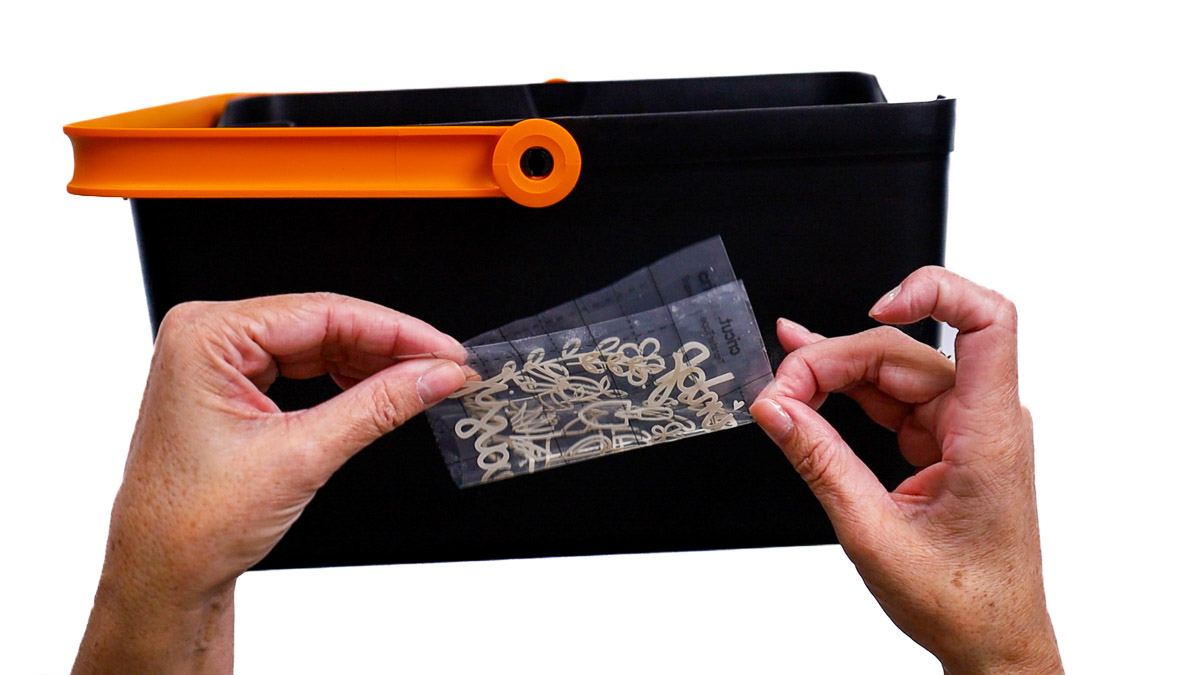 Hands hold a small vinyl decal with transfer tape in front of a black garden bucket, lining it up to find the center before applying.